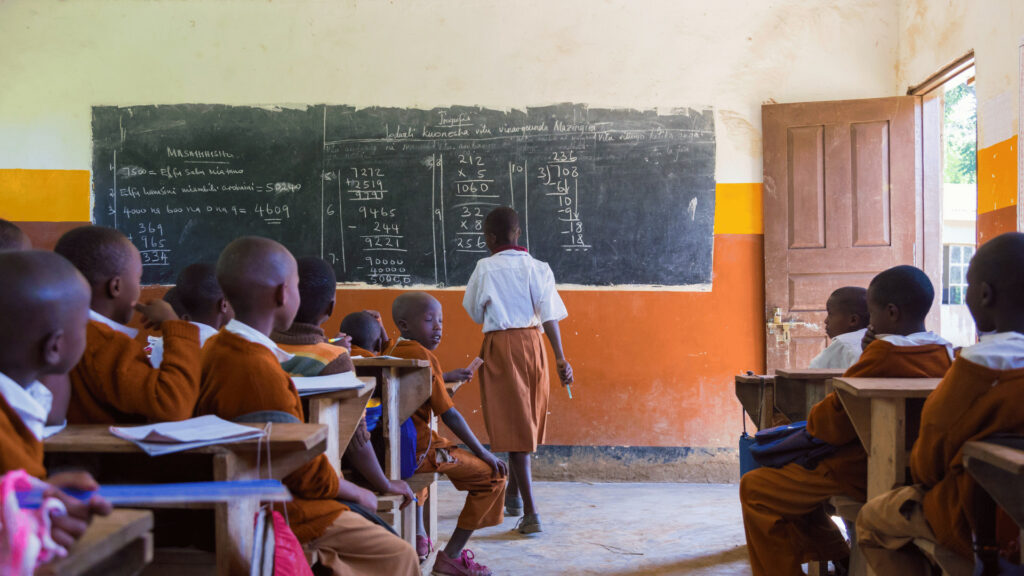 Students in a rural African classroom engaged in a lesson with a teacher at the chalkboard.